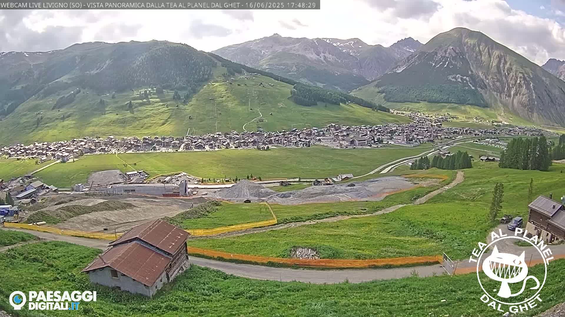 A mountain village nestled in a valley, with construction visible in the foreground, under a partly cloudy sky.