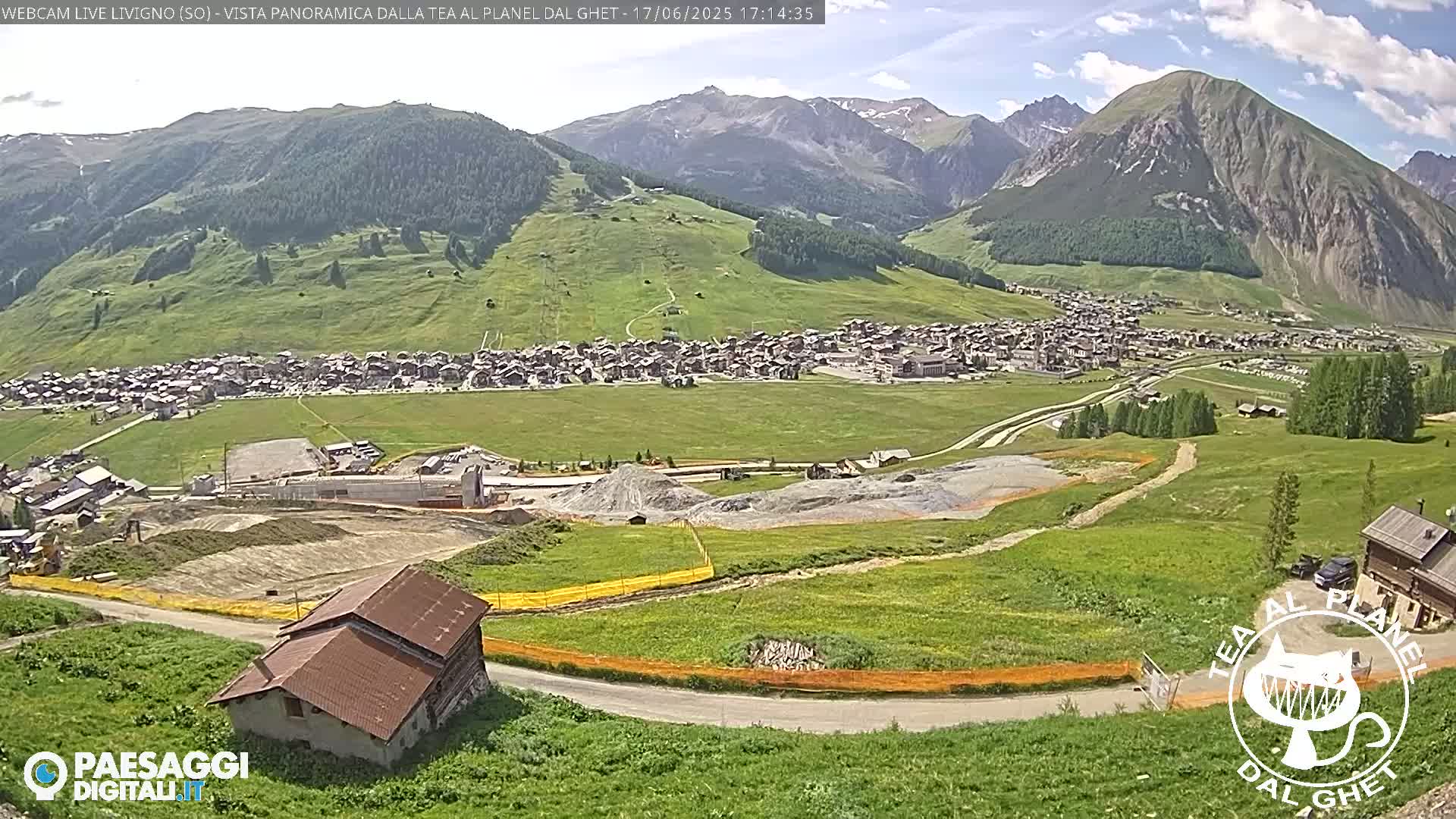 A mountain village nestled in a valley, with green hills and a partly cloudy sky, is viewed from an elevated vantage point, showing construction activity in the foreground.
