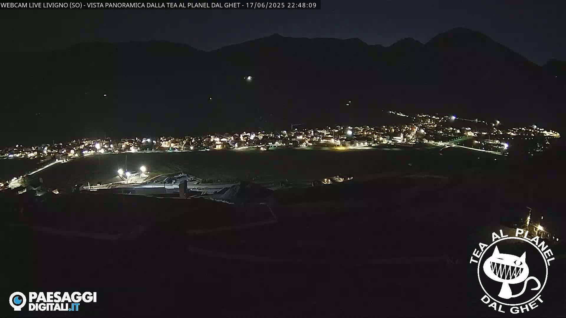 A nighttime, panoramic view of a small town nestled in a valley, illuminated by lights, under a dark sky.