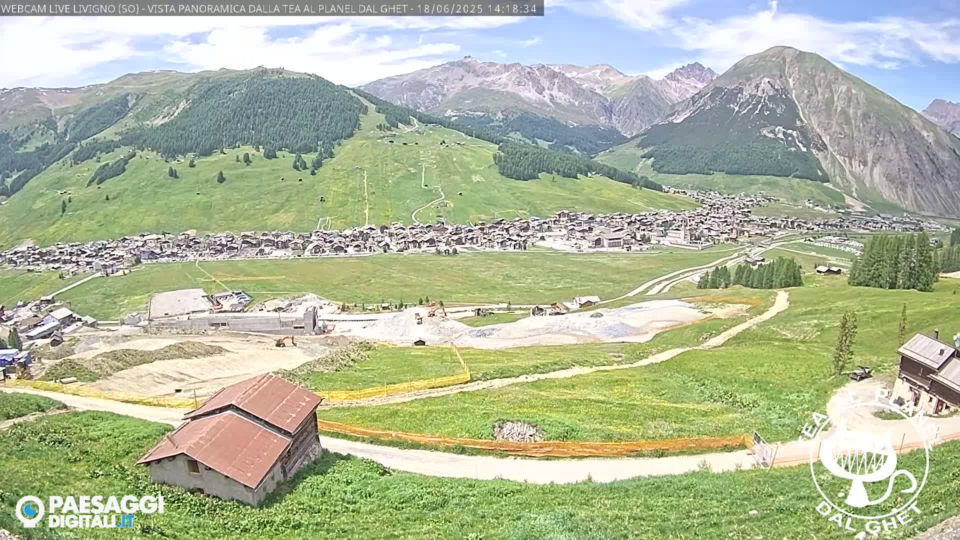 A sunny day reveals a mountain village nestled in a green valley, with construction visible in the foreground and mountains in the background.