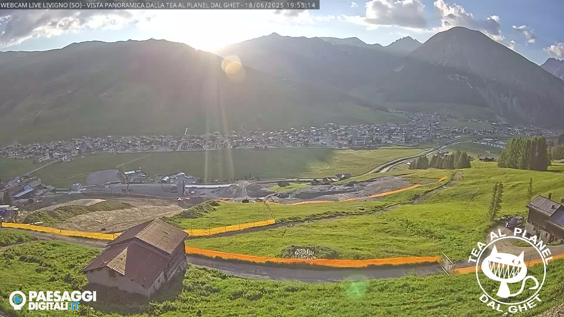 A sunlit panoramic view of a mountain town nestled in a valley, with grassy hills and a small building in the foreground, under partly cloudy skies.