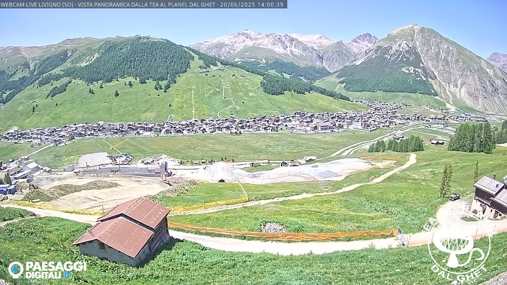 A sunny, high-altitude view of a village nestled in a valley surrounded by green hills and snow-capped mountains.