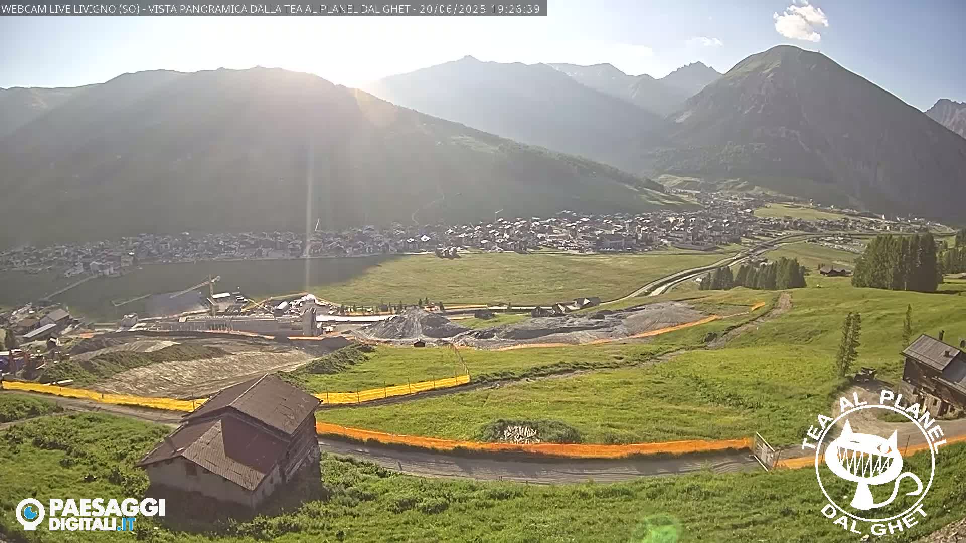 A sunny, high-angle view shows a small town nestled in a valley between mountains, with construction visible in the foreground and green fields extending towards the mountains.