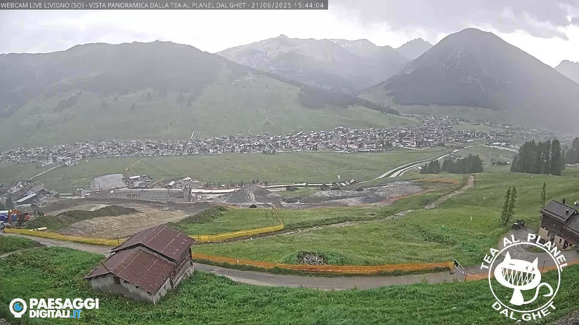 A panoramic view shows a town nestled in a valley between mountains on an overcast day with light rain.