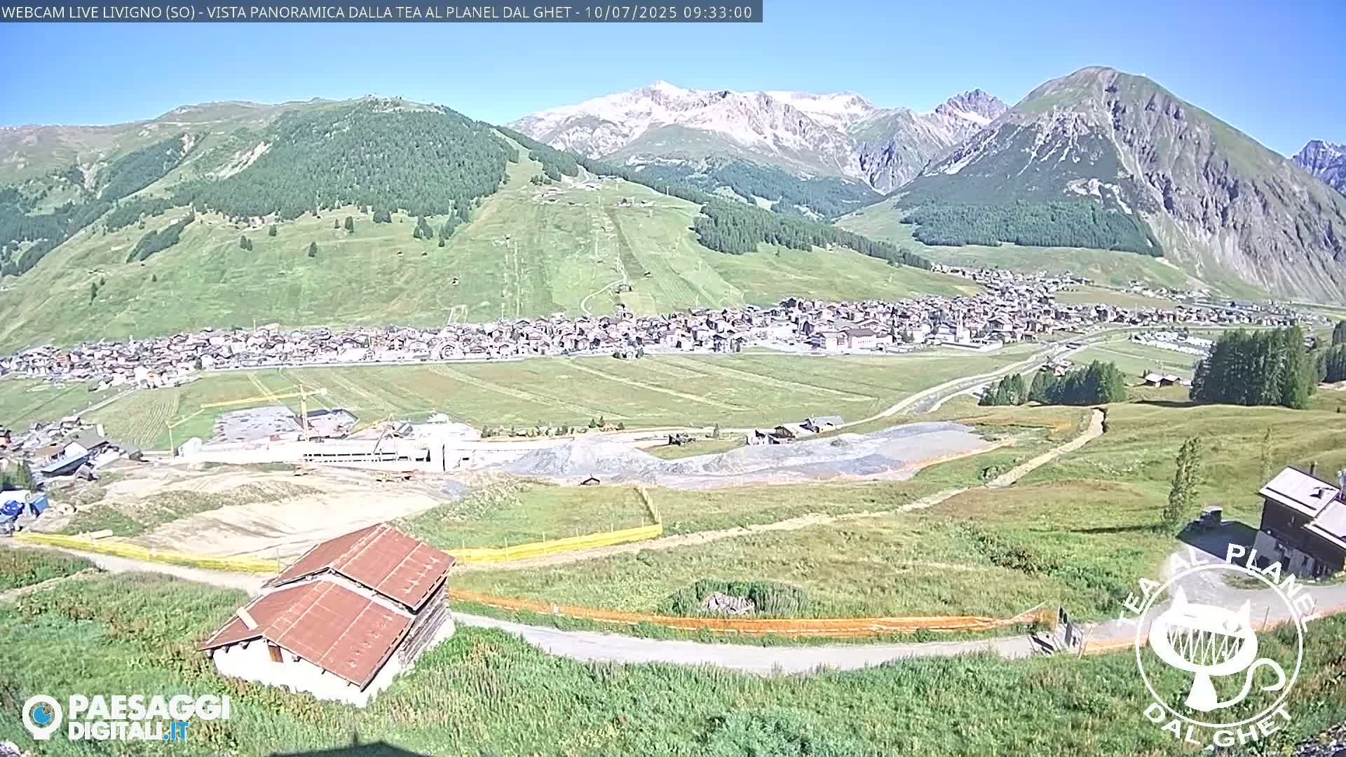 A sunny mountain valley town is visible, with construction taking place in the foreground, and snow-capped mountains in the background.