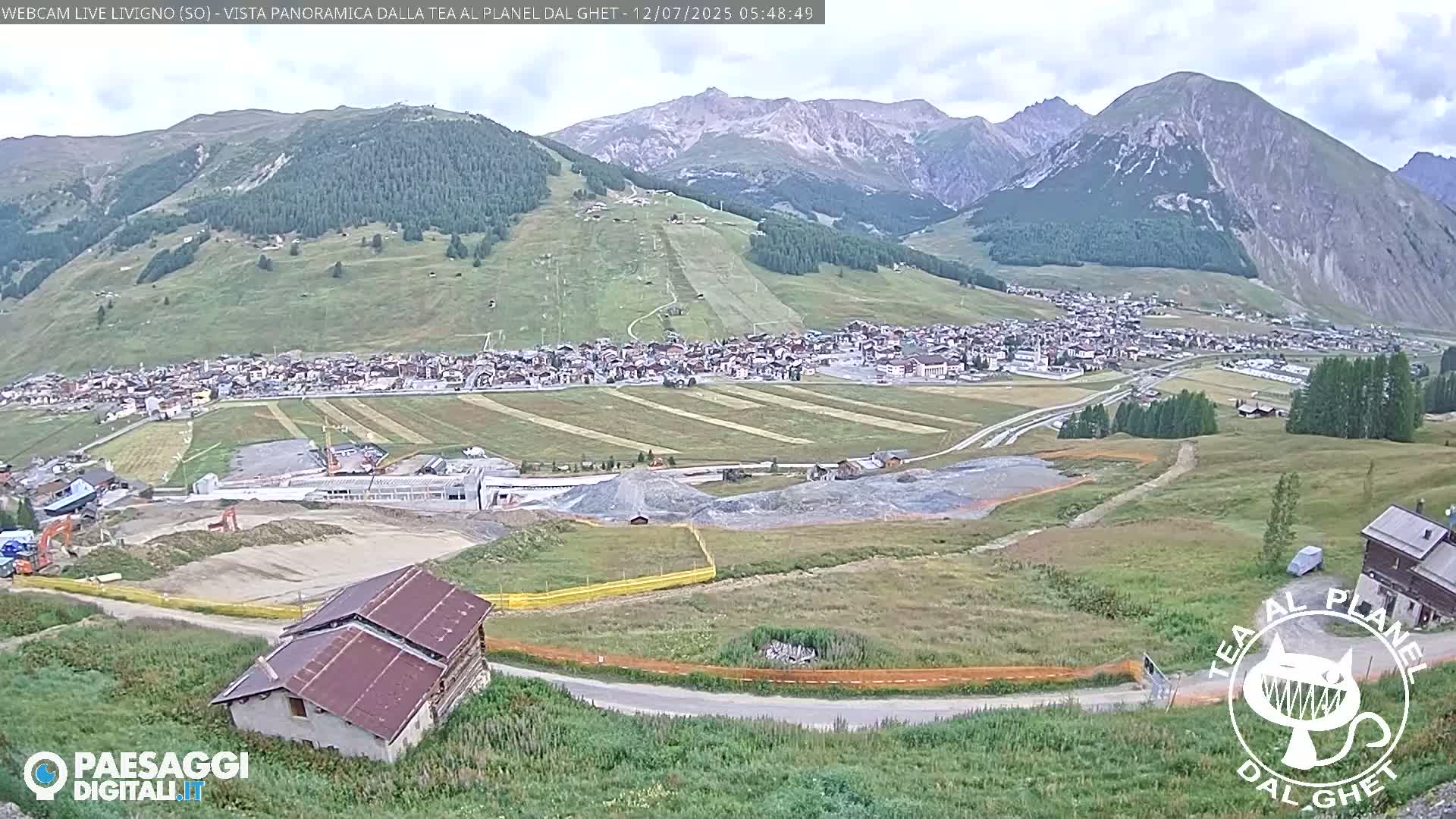 A sunny mountain valley town is visible, with construction taking place in the foreground, and snow-capped mountains in the background.