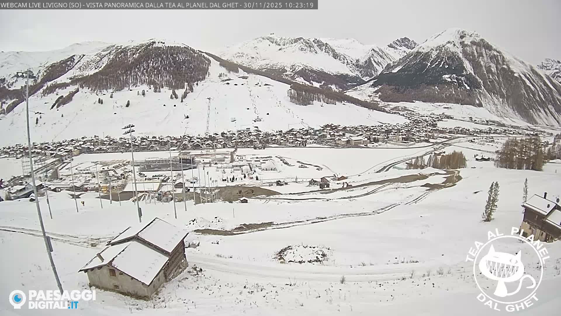 A wide panoramic view captures a snow-covered alpine valley and village under an overcast sky, with majestic snow-capped mountains dominating the background and ski slopes visible on their flanks.