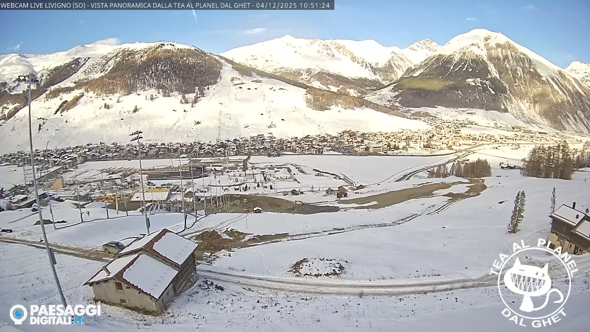A panoramic view captures a snow-covered mountain valley with a village nestled along a partially frozen river, under a clear, bright blue sky.