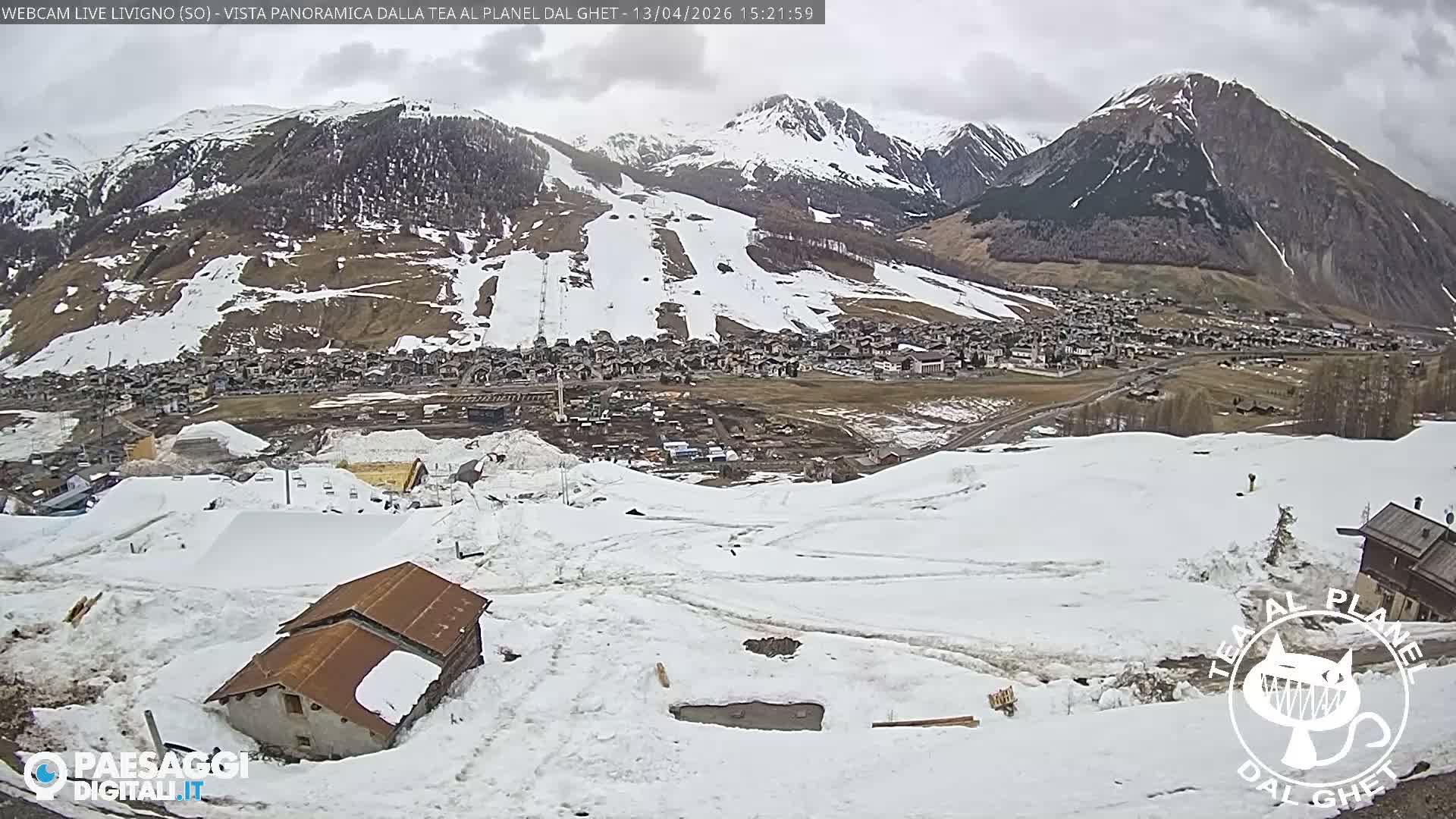 A panoramic view captures a snow-covered mountain valley with a village nestled along a partially frozen river, under a clear, bright blue sky.
