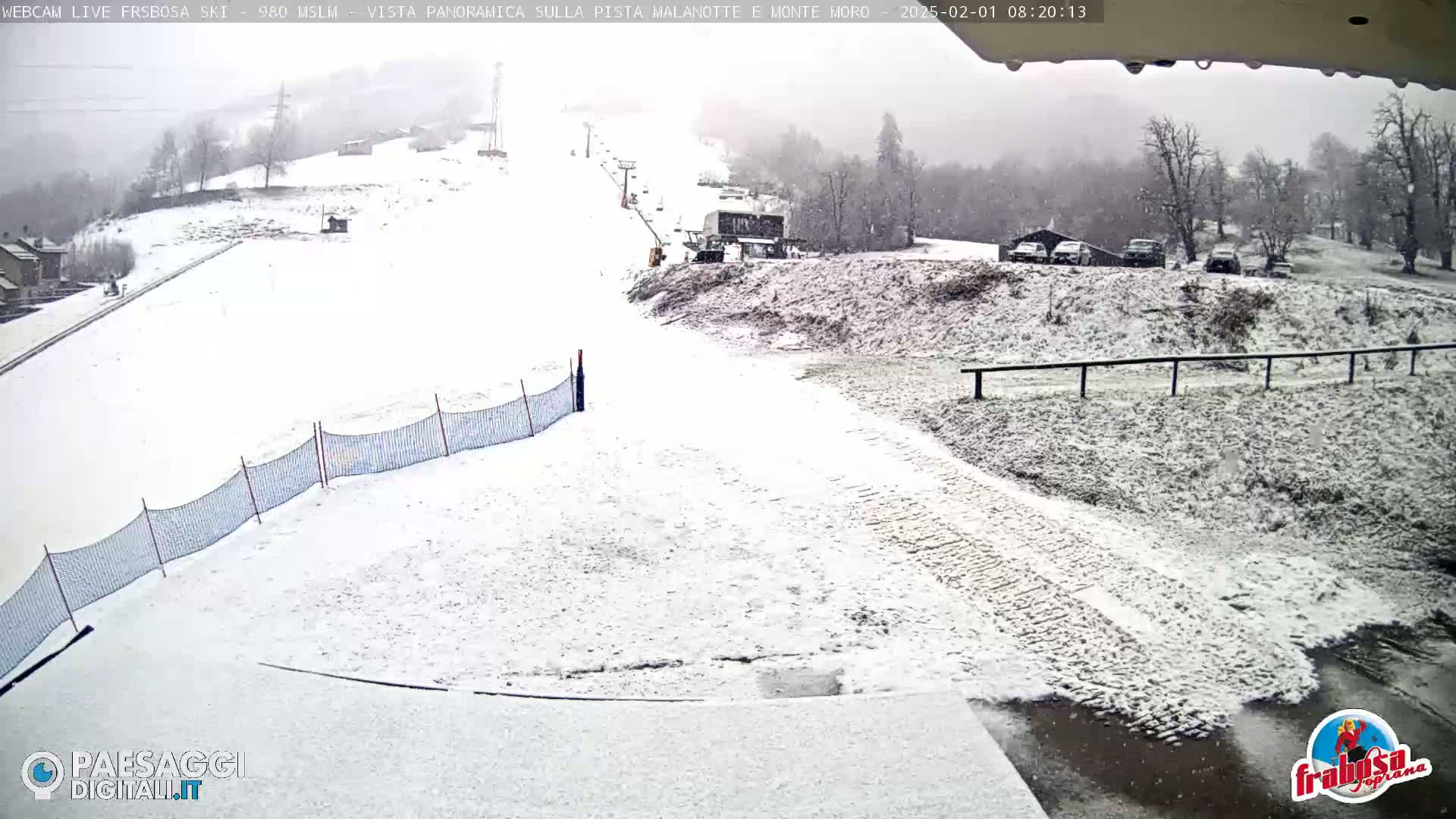 A snow-covered ski slope with a ski lift and several cars parked nearby during a light snowfall.