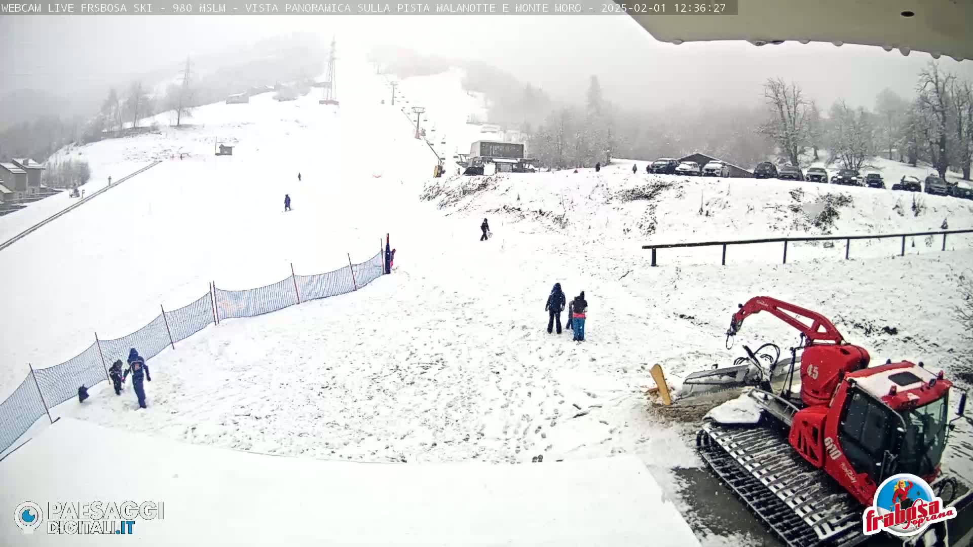 A snow-covered ski slope with several people and a red snow groomer, under overcast, snowy conditions.