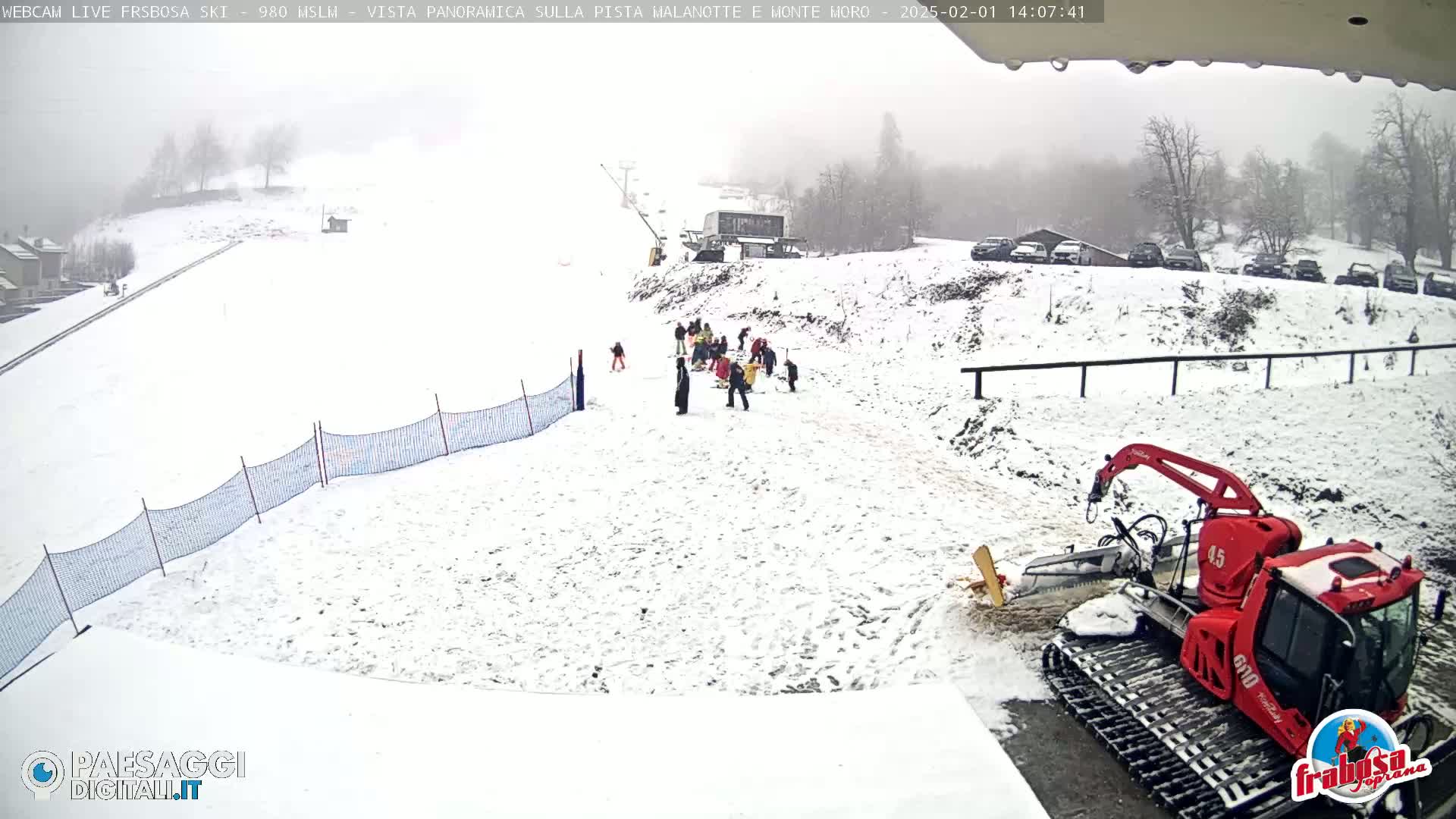 A snow-covered ski slope with several people and a red snow groomer, under an overcast sky.