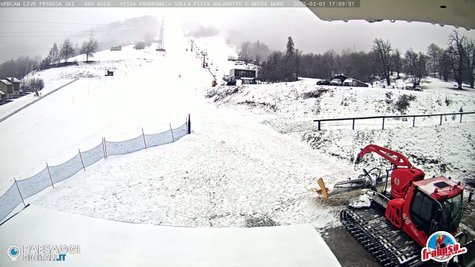 A snow-covered ski slope with a ski lift and a red snow groomer parked at the bottom, under an overcast sky.