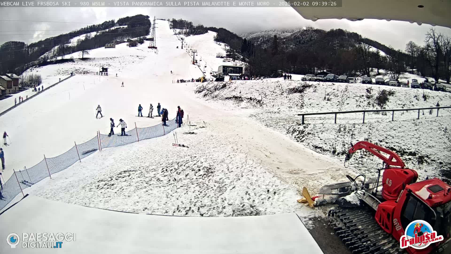 A snow-covered ski slope with several skiers and a snow groomer, under an overcast sky.