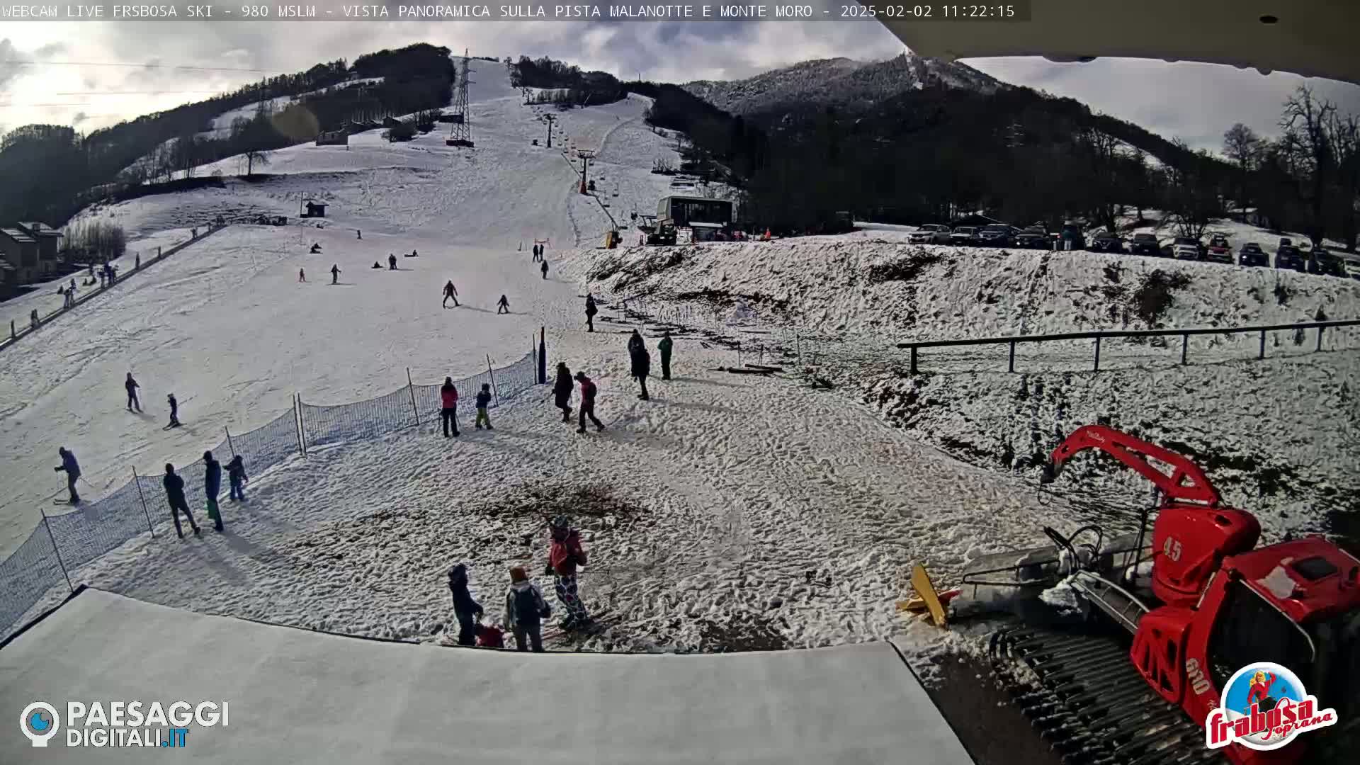 A snow-covered ski slope with several people skiing and snowboarding, a snow groomer, and a parking lot in the background under a partly cloudy sky.