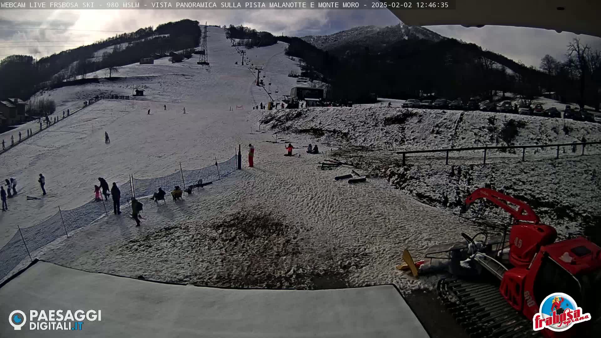 A snow-covered ski slope with several people skiing and standing around, a ski lift, and a snow groomer in the foreground under a partly cloudy sky.