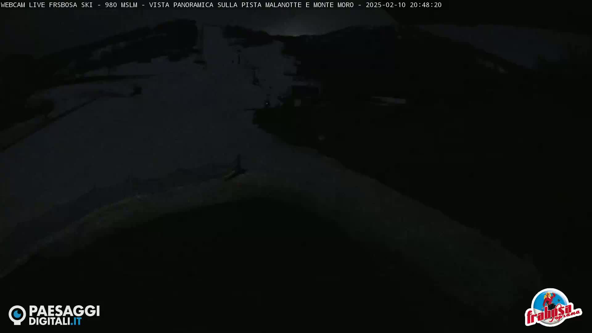 A dark, snowy, nighttime scene shows a mostly empty ski slope.