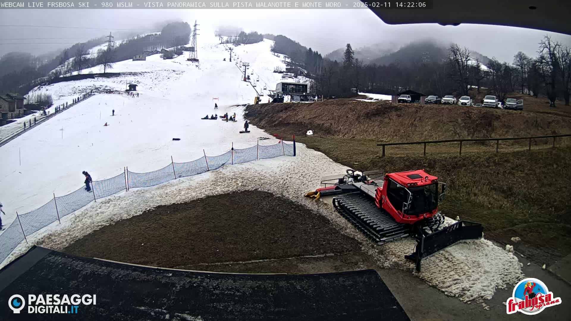 A snowcat is parked near a snowy ski slope with several people visible, under an overcast sky.
