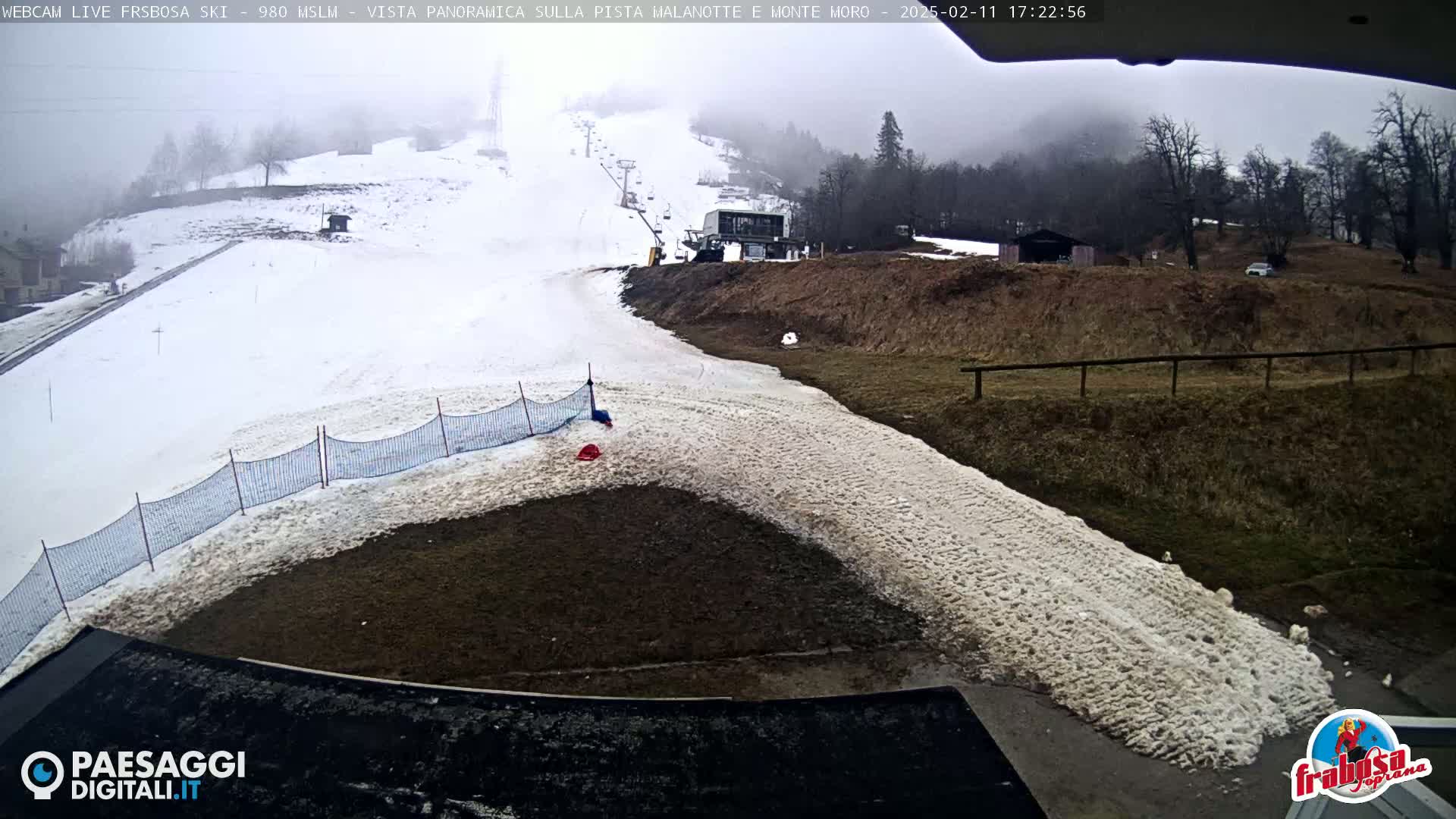 A foggy view of a snow-covered ski slope with a chairlift and a small building, partially melted snow at the bottom of the slope, and a wire fence.