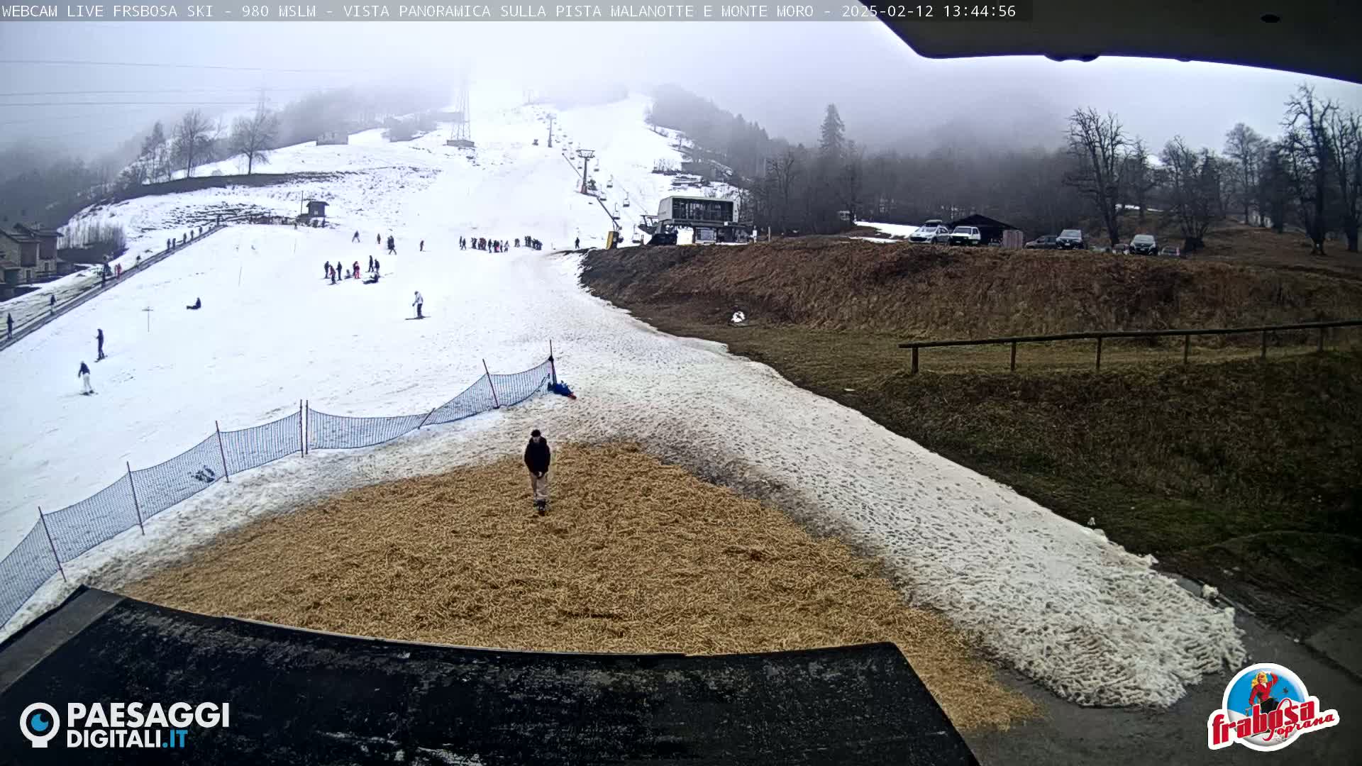 A snowy ski slope with a few skiers and a person walking on a tan surface below, viewed on an overcast day.