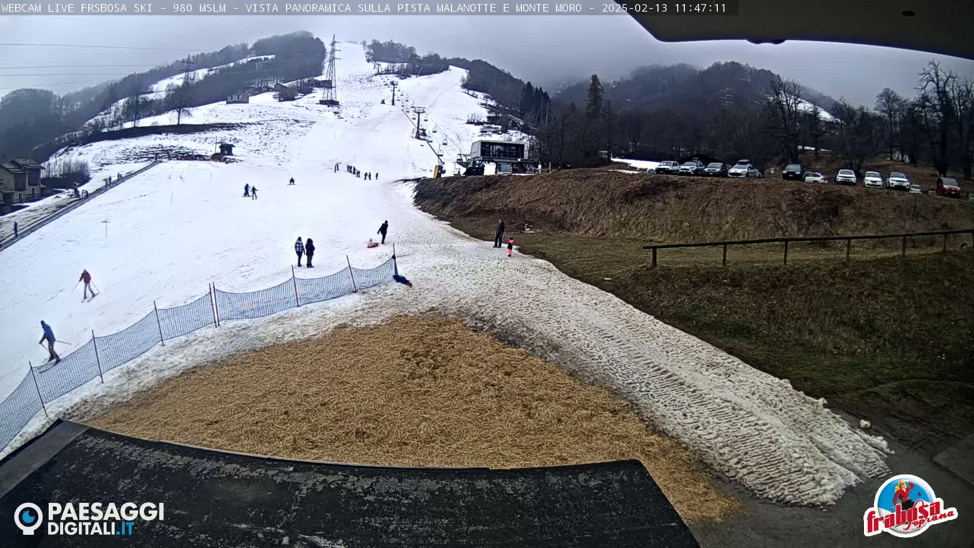 A snowy ski slope with a few skiers and people on foot, a ski lift, and a parking lot of cars in the background under an overcast sky.