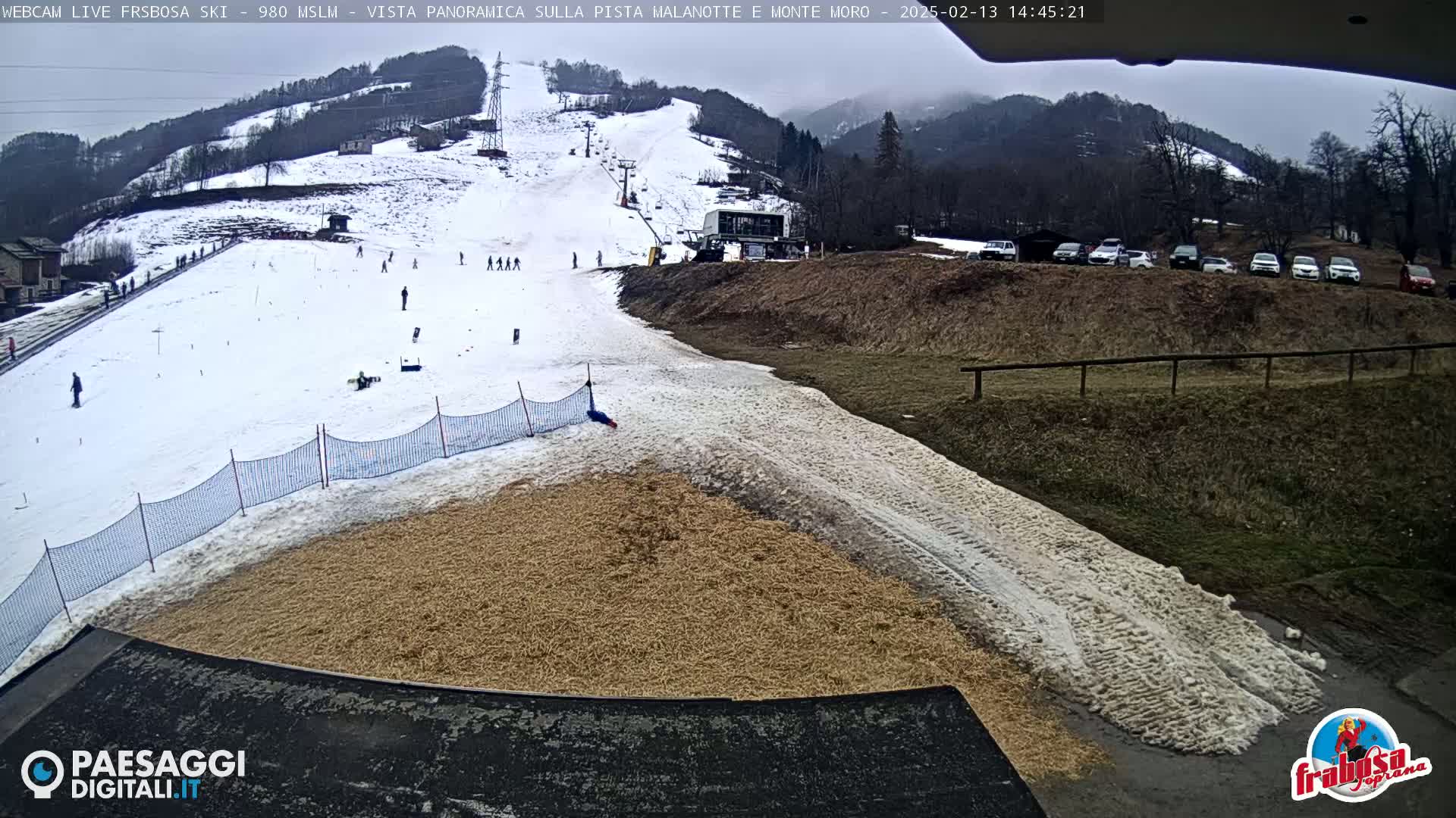 A snowy ski slope with several skiers, a ski lift, and a parking lot of cars in the background under an overcast sky.