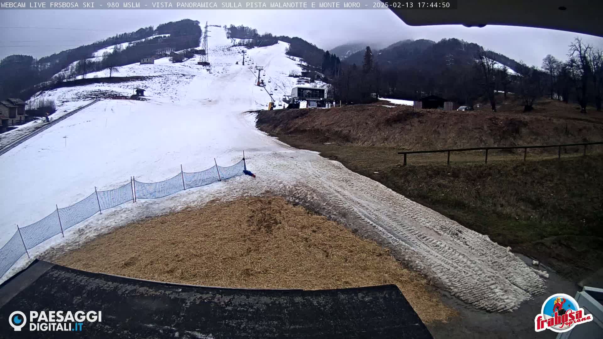 A snowy ski slope with a chairlift and a hay-covered area at the bottom, under an overcast sky.
