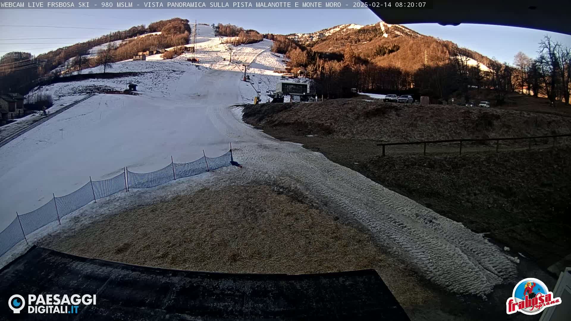A snow-covered ski slope is viewed from a low angle on a sunny day, with a small building and several cars visible in the background.
