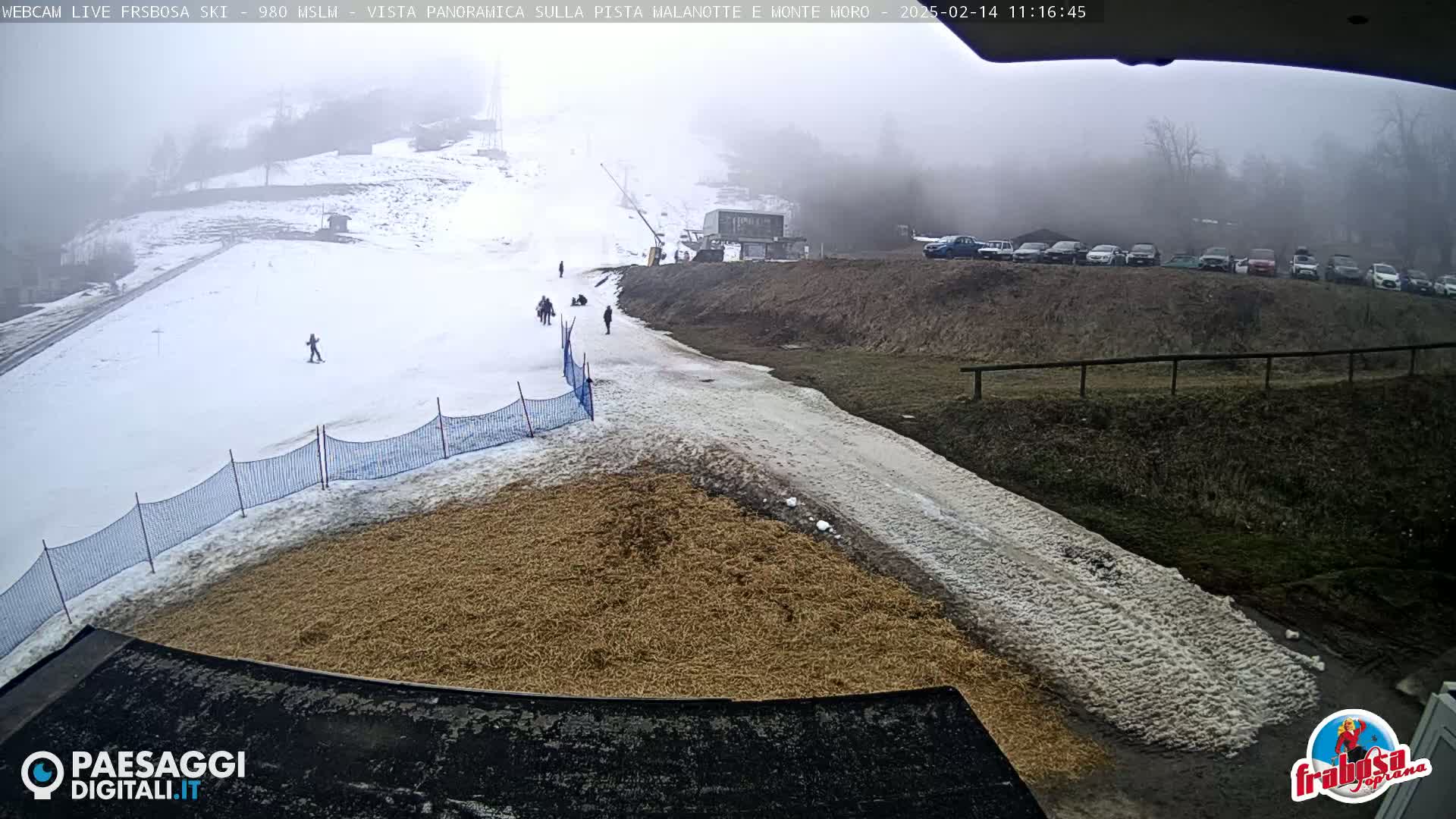 A foggy snowy ski slope with a few people skiing and a parking lot full of cars in the background.