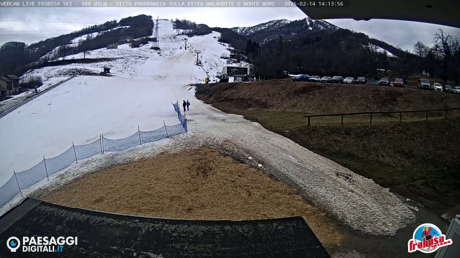 A snowy ski slope with a ski lift and several cars parked in a lot nearby, under an overcast sky.