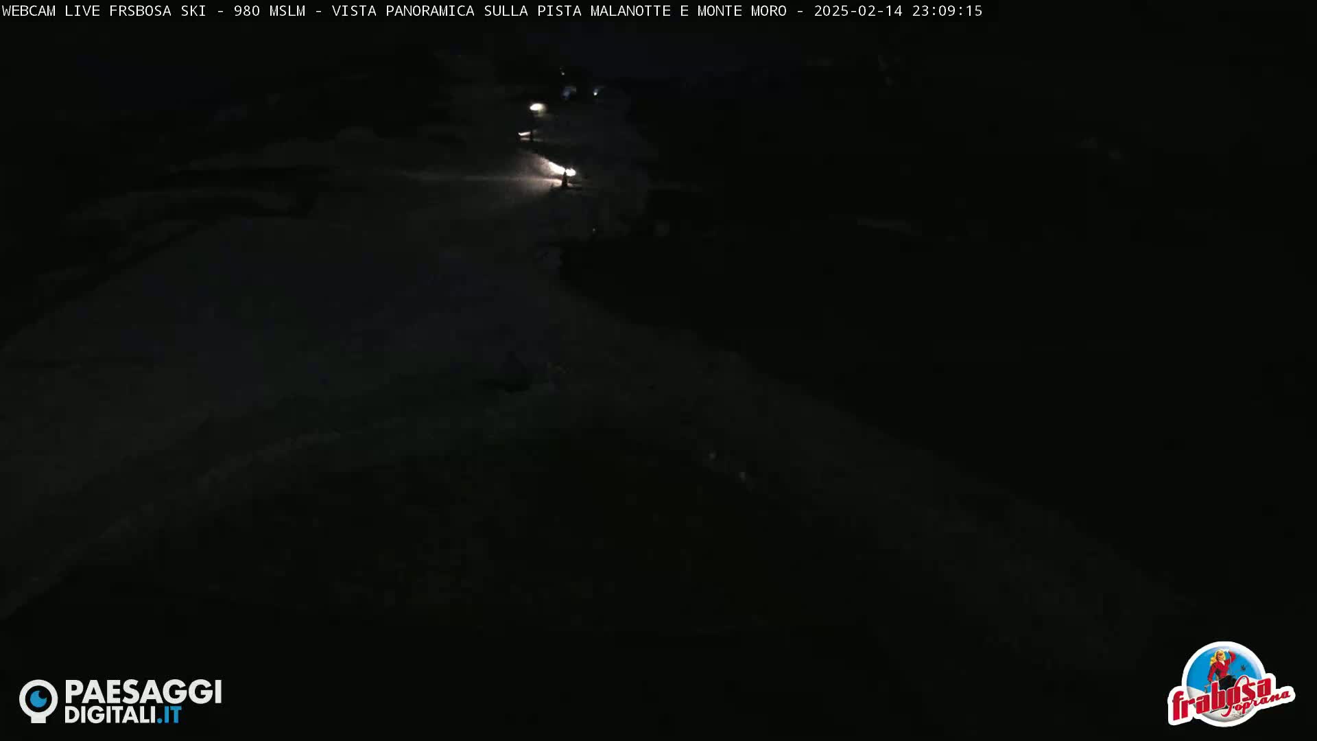 A dark, nighttime view of a snow-covered slope, illuminated by a few distant lights.