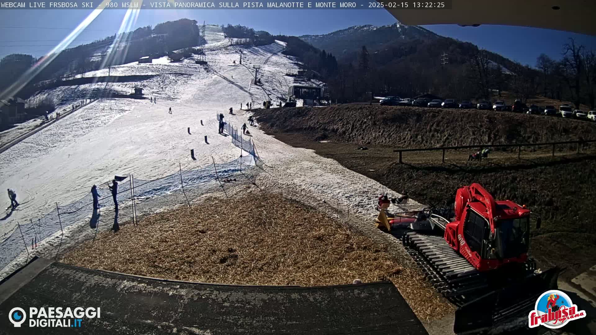 A sunny day at a ski resort shows a snow-covered slope with several people skiing and snowboarding, a snow groomer in the foreground, and a parking lot filled with cars in the background.