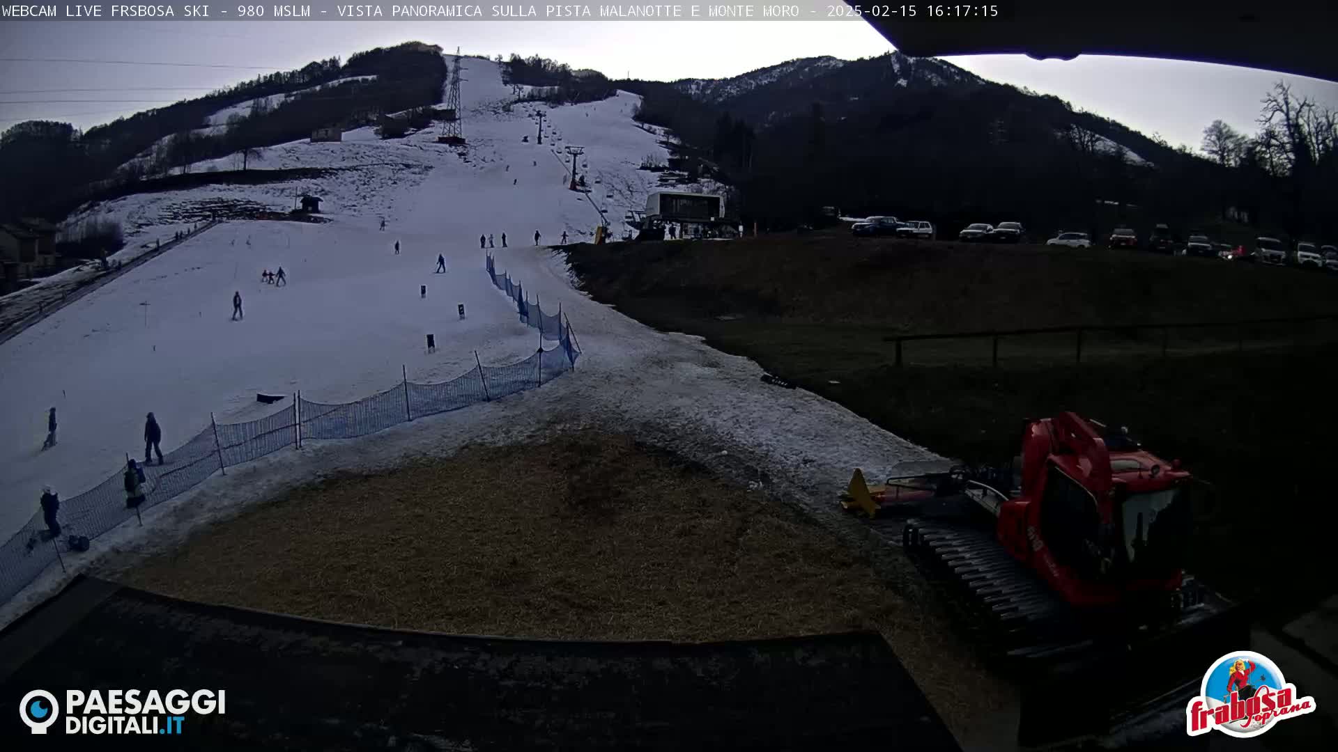 A snow-covered ski slope with several skiers, a snow groomer, and a parking lot full of cars under an overcast sky.