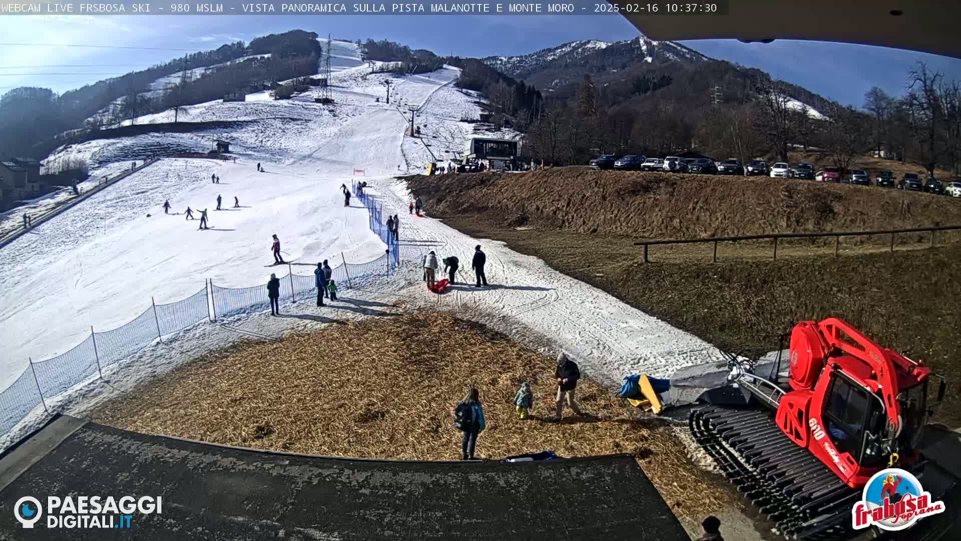 A snowy ski slope with several people skiing and others standing around, a snow groomer at the bottom, and a sunny sky above.
