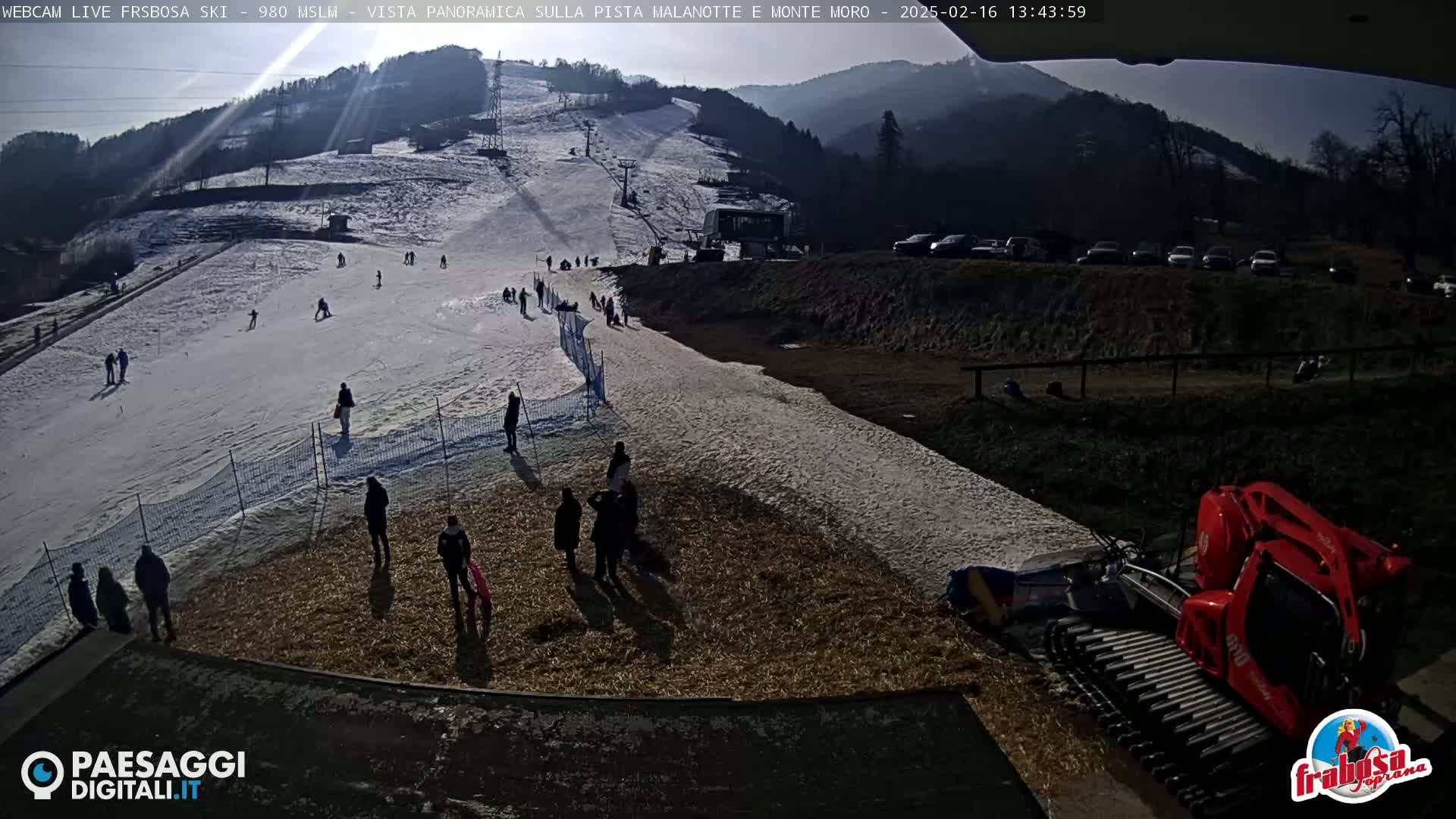A sunny day at a snow-covered ski slope shows several people skiing and standing around, with a snow groomer parked nearby.