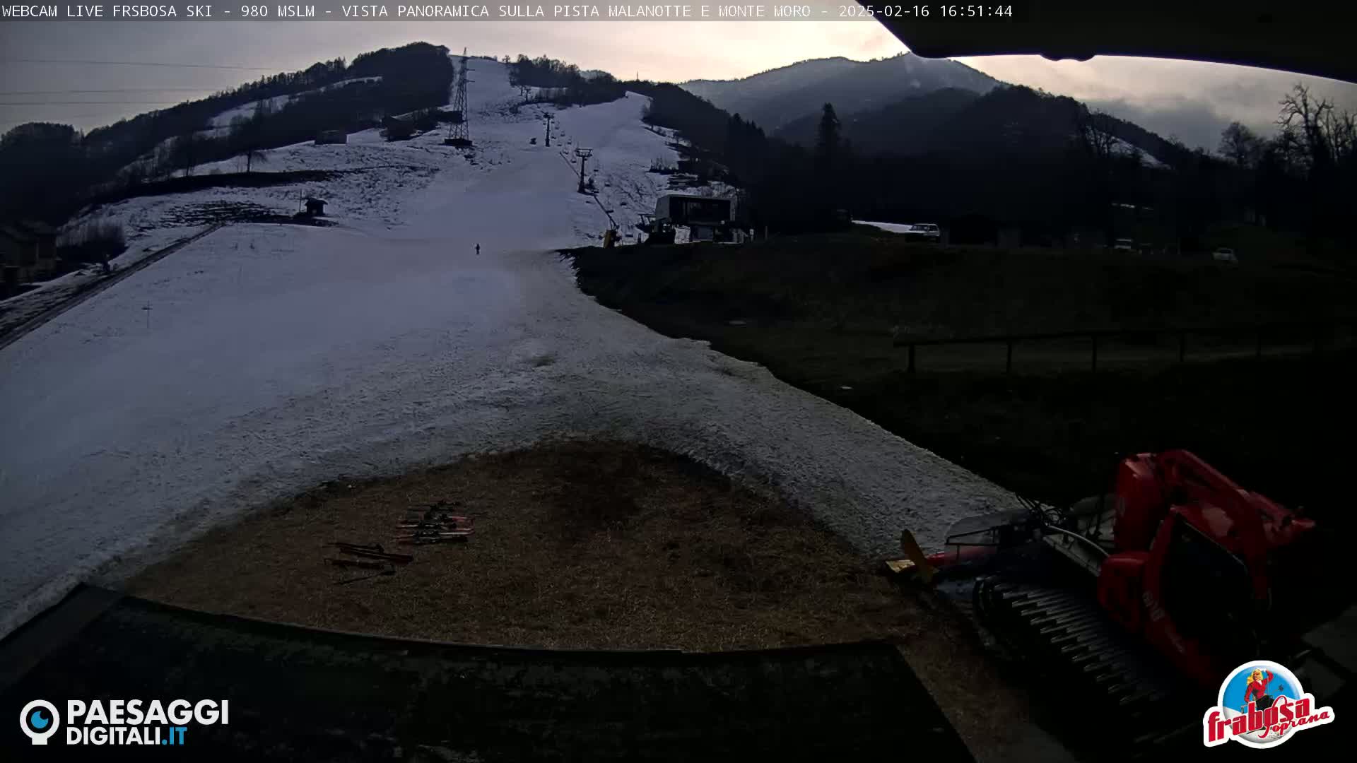 A snow-covered ski slope at dusk, with a snowcat parked at the bottom and a few pairs of skis lying on the ground.