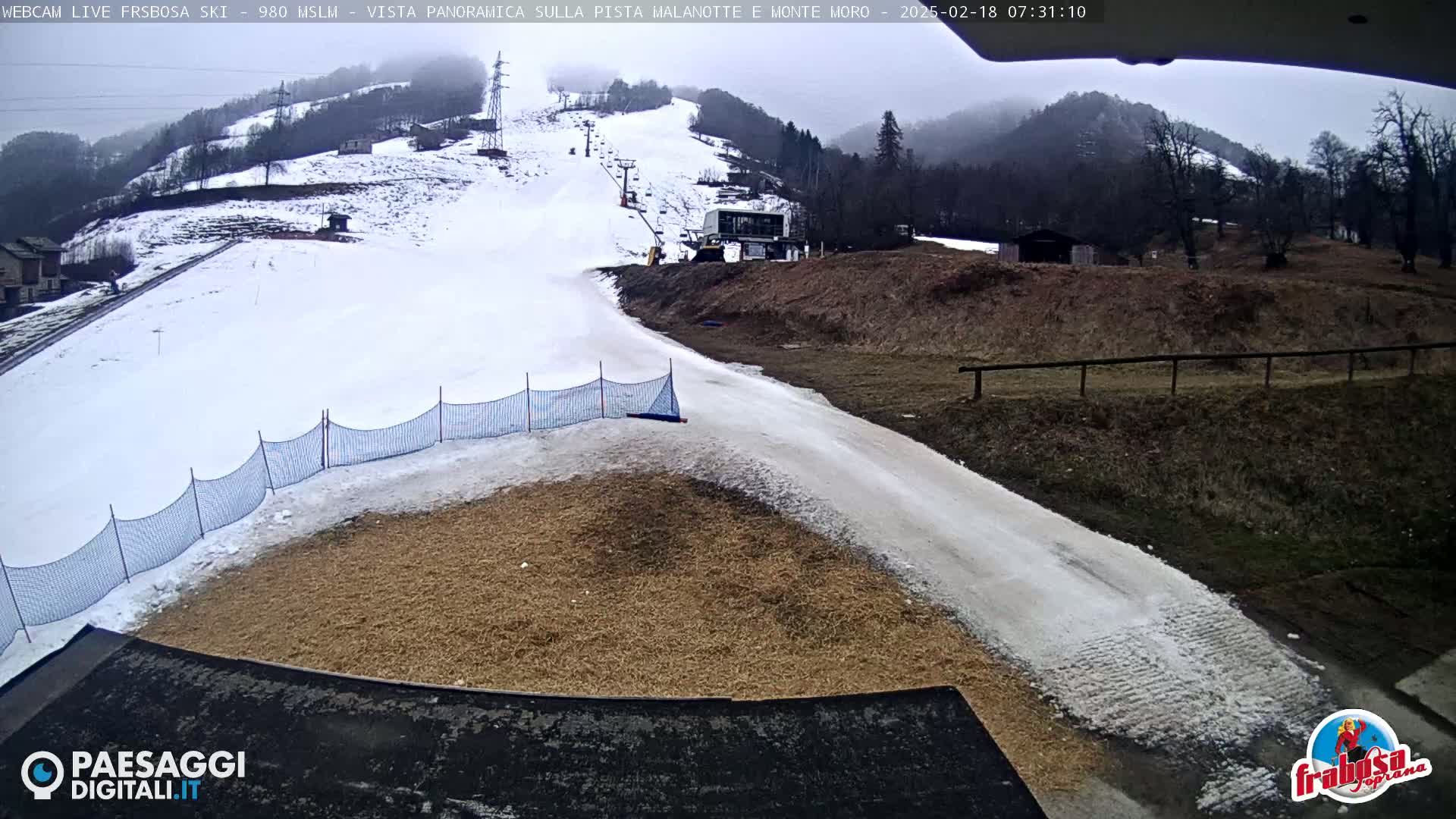 A snow-covered ski slope with a ski lift and a small area of bare ground at the bottom, under an overcast sky.