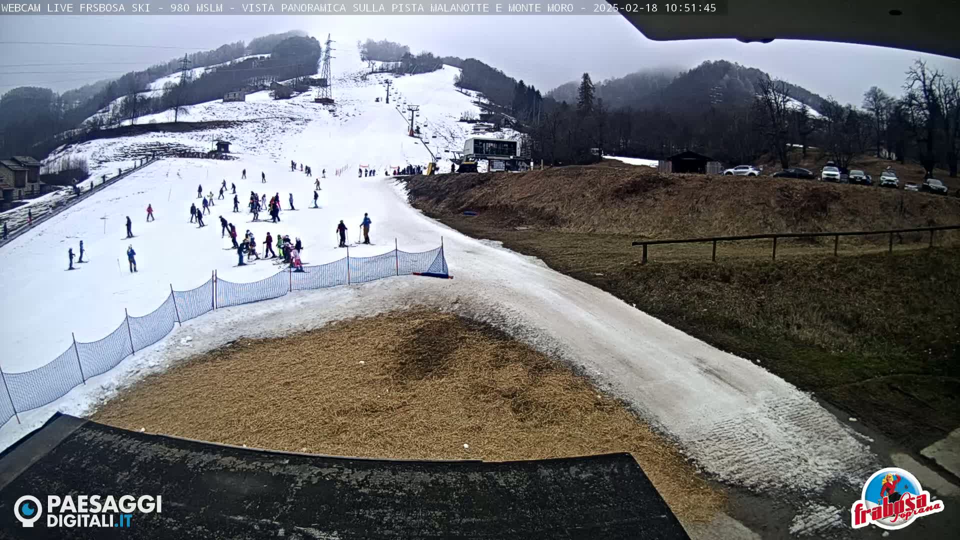 A snowy ski slope with skiers and snowboarders, a ski lift, and parked cars in the background under an overcast sky.