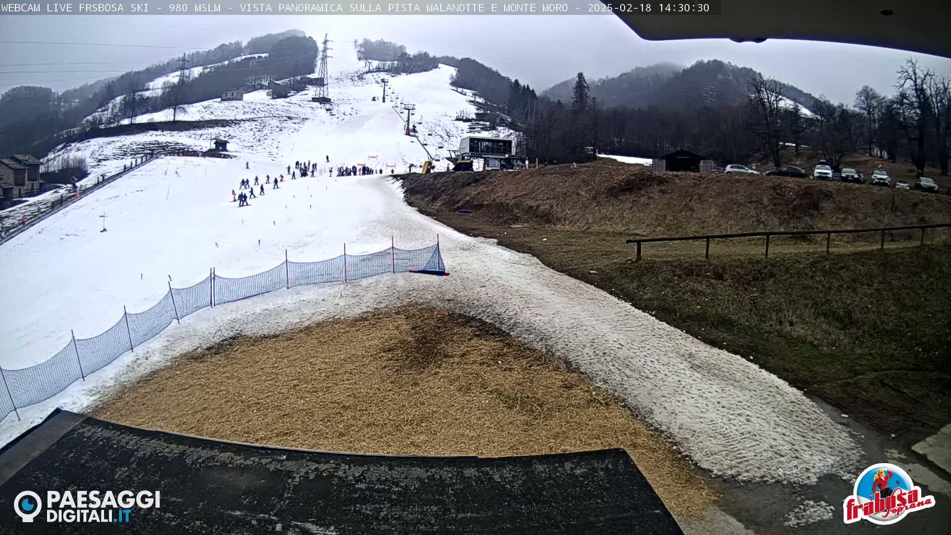 A snowy ski slope with a few skiers visible, a lift, and a lower section with a tan surface next to a snow-covered path; the overcast sky suggests a cloudy day.