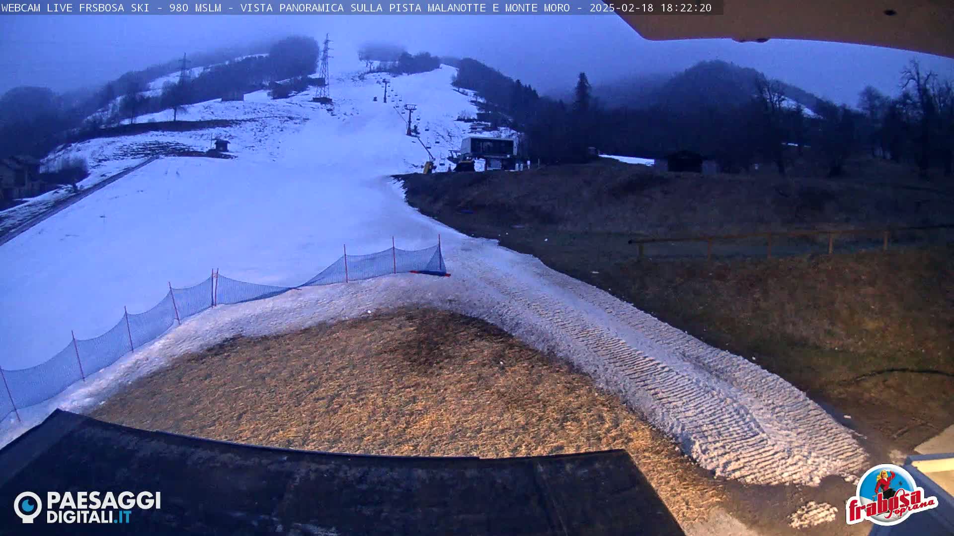 A snow-covered ski slope at dusk, with a section of bare ground and a safety net at the bottom.