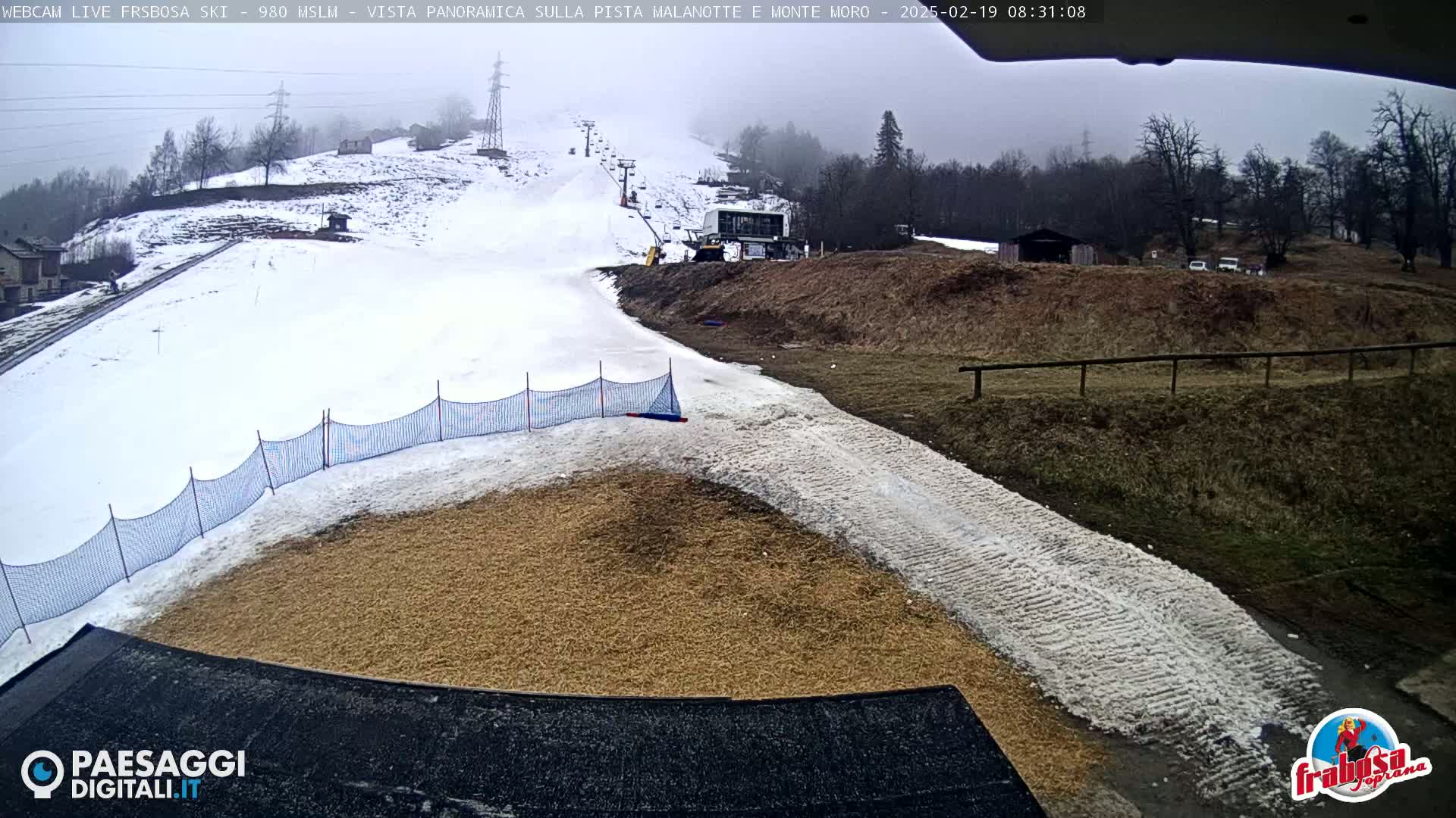 A snowy ski slope with a ski lift and a snow-covered landing area next to a tan, granulated surface, all under an overcast sky.