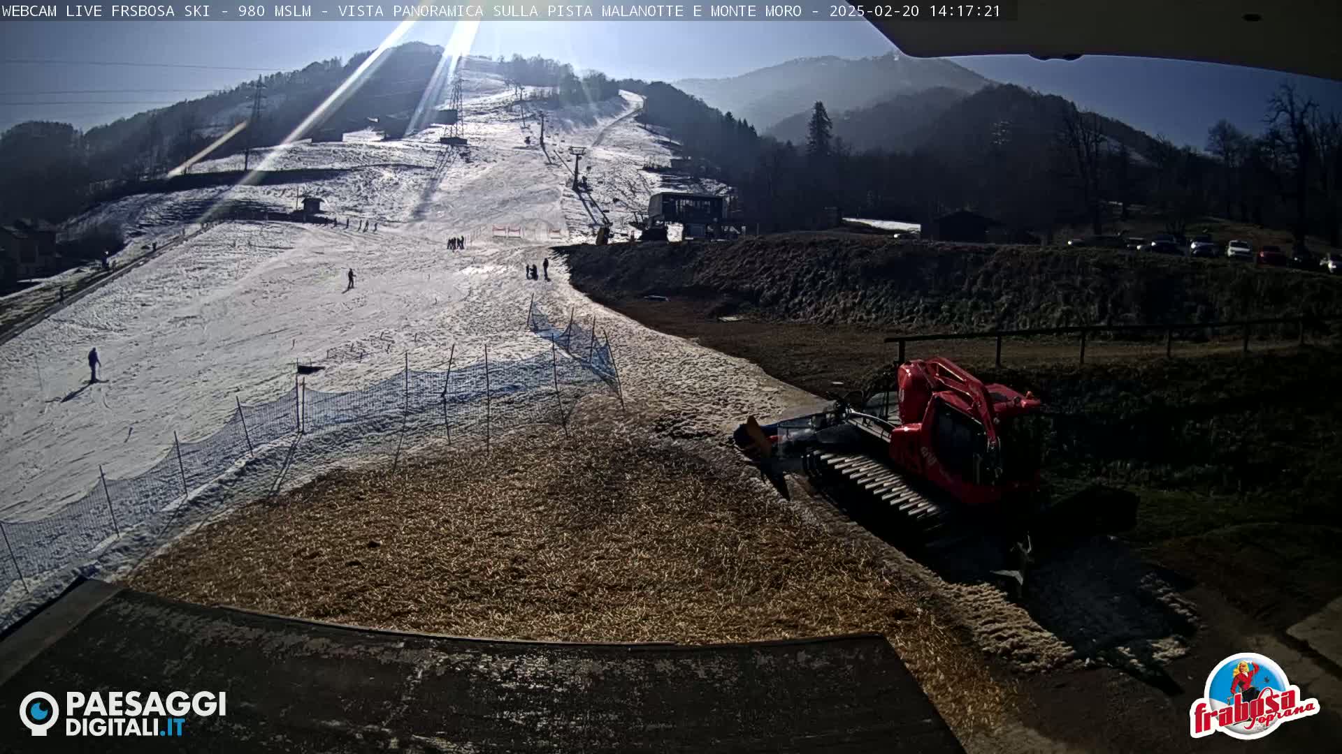 A sunny, snow-covered ski slope with a few skiers, a snow groomer, and a parking lot in the background.