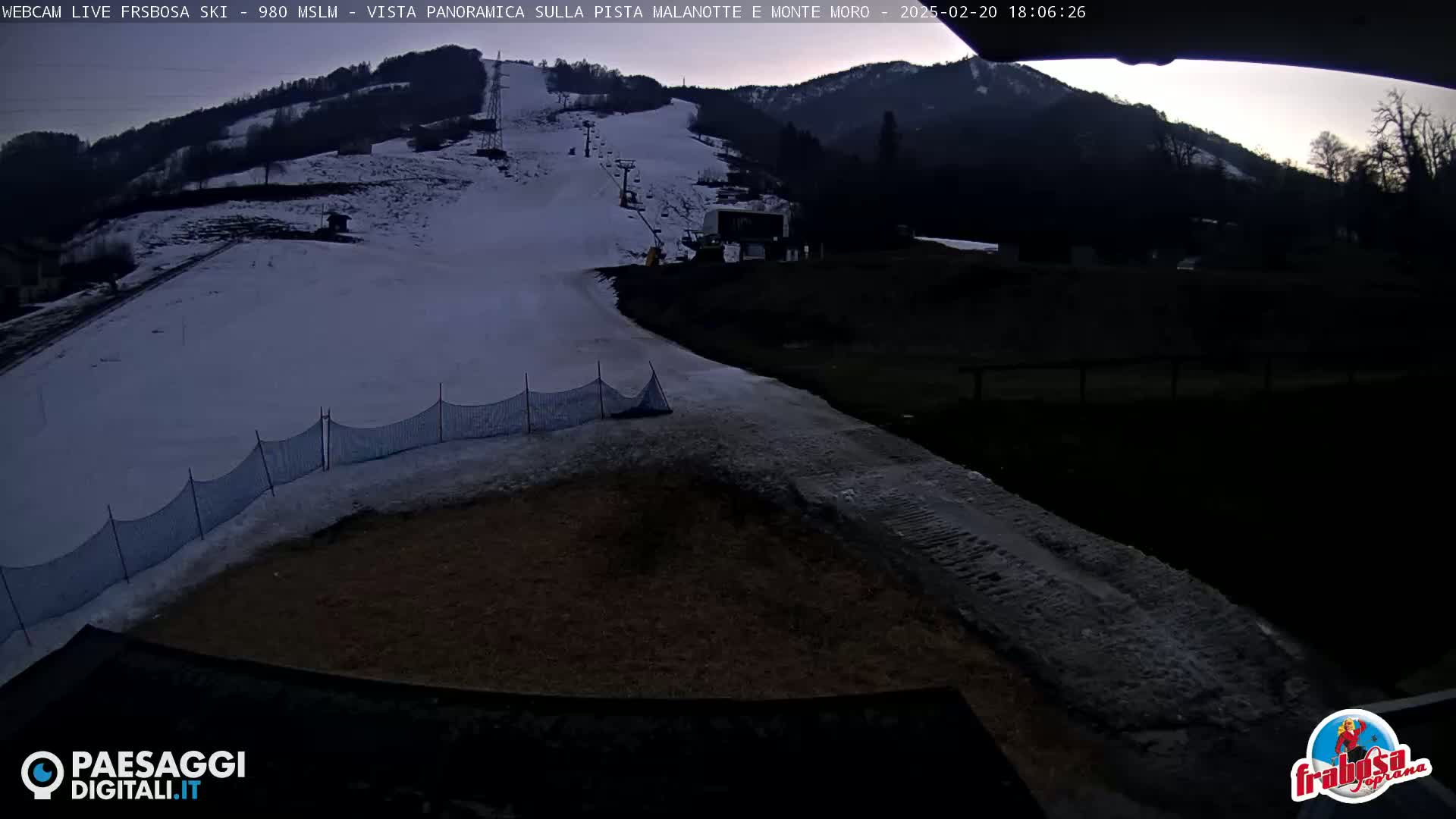 A snow-covered ski slope at dusk, with a ski lift visible in the background and a section of netting near the bottom.