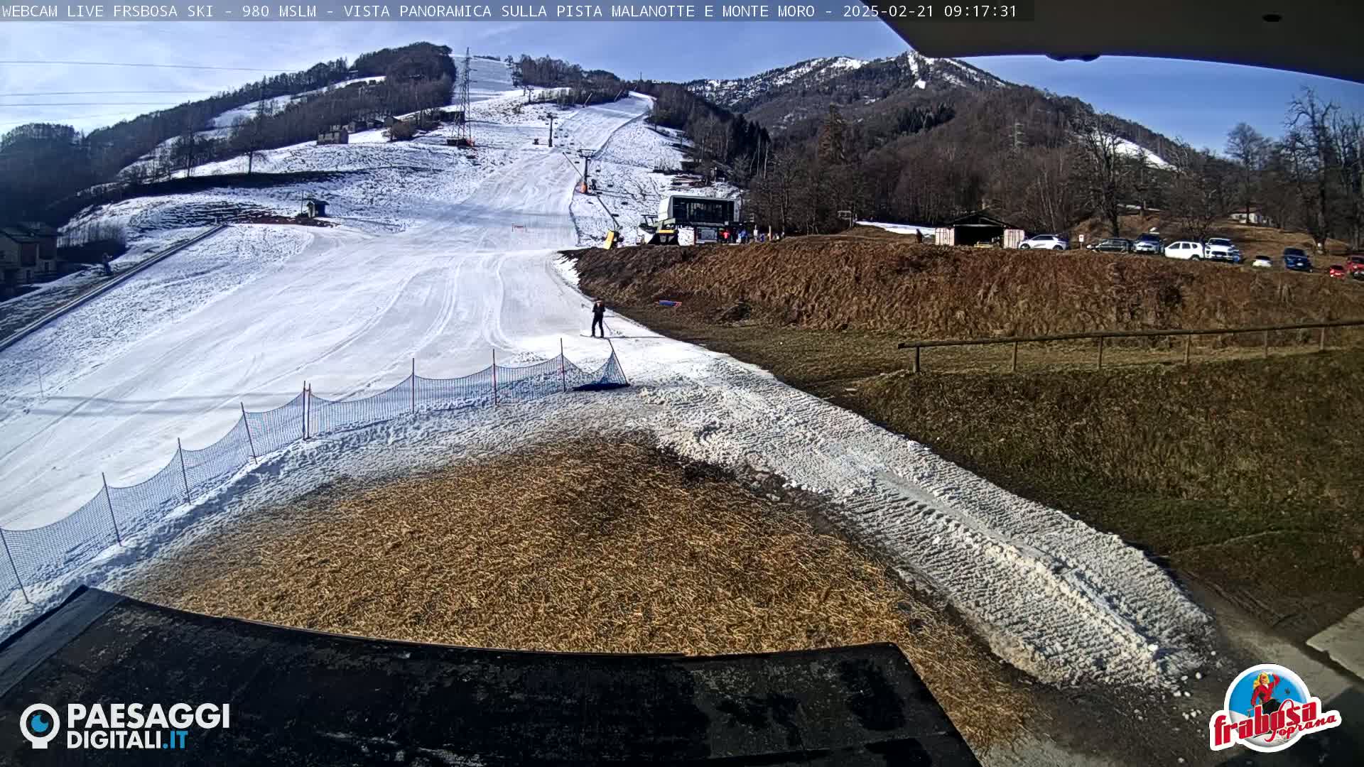 A snowy ski slope with a person skiing, a ski lift, and a parking lot with cars visible in the background under a sunny sky.