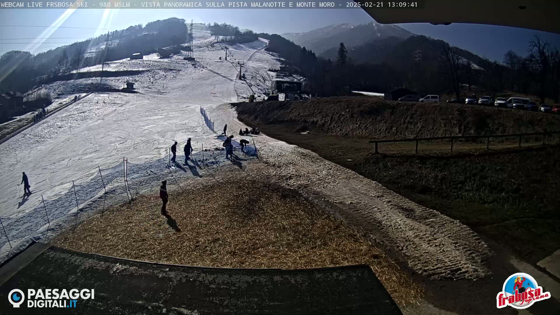 A sunny day at a ski resort shows a snow-covered ski slope with several people skiing and standing around, alongside a dirt area with some cars parked nearby, and a mountain in the background.