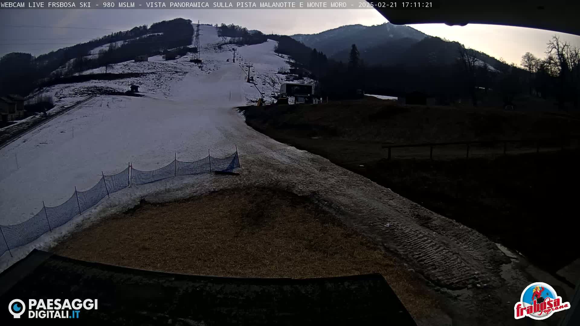 A snow-covered ski slope with a partially snow-covered access road is visible under a partly cloudy sky at dusk.