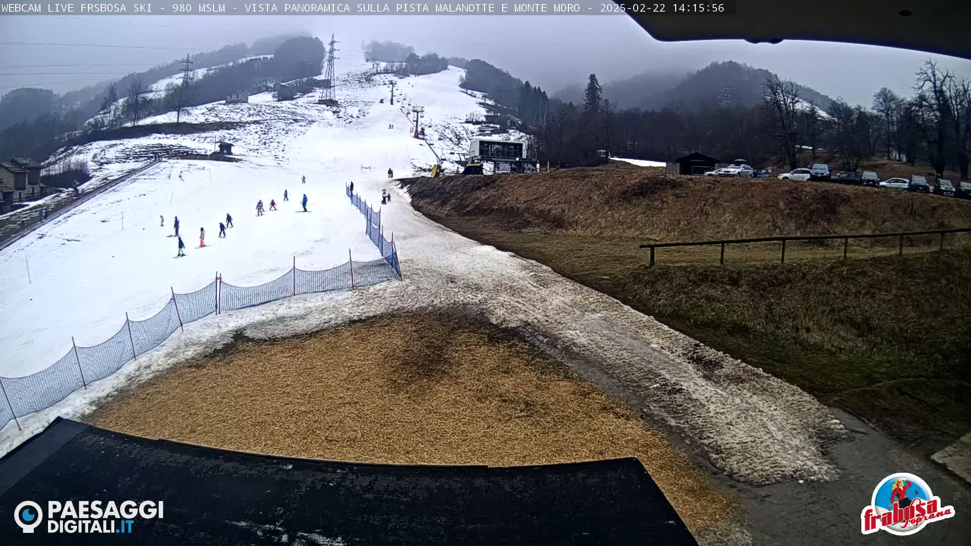 A snowy ski slope with several skiers, a ski lift, and a parking lot with cars in the background on an overcast day.