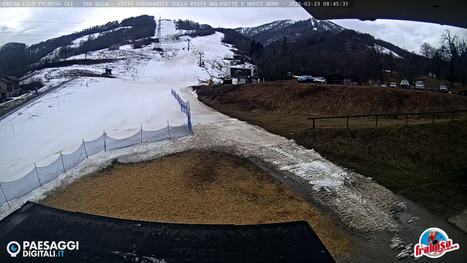 A mostly snow-covered ski slope with a ski lift and several cars parked nearby, under a cloudy sky.