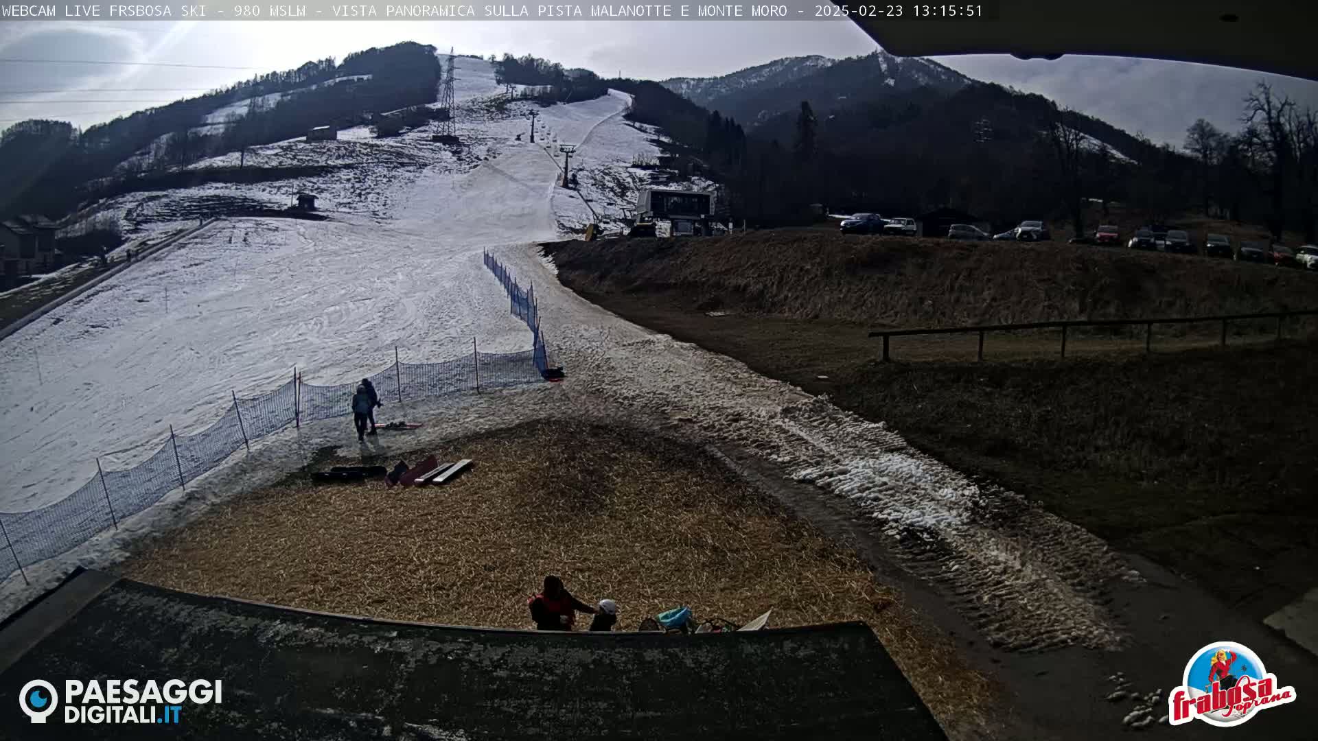 A snow-covered ski slope with a chairlift and several people near the bottom, under a partly cloudy sky.