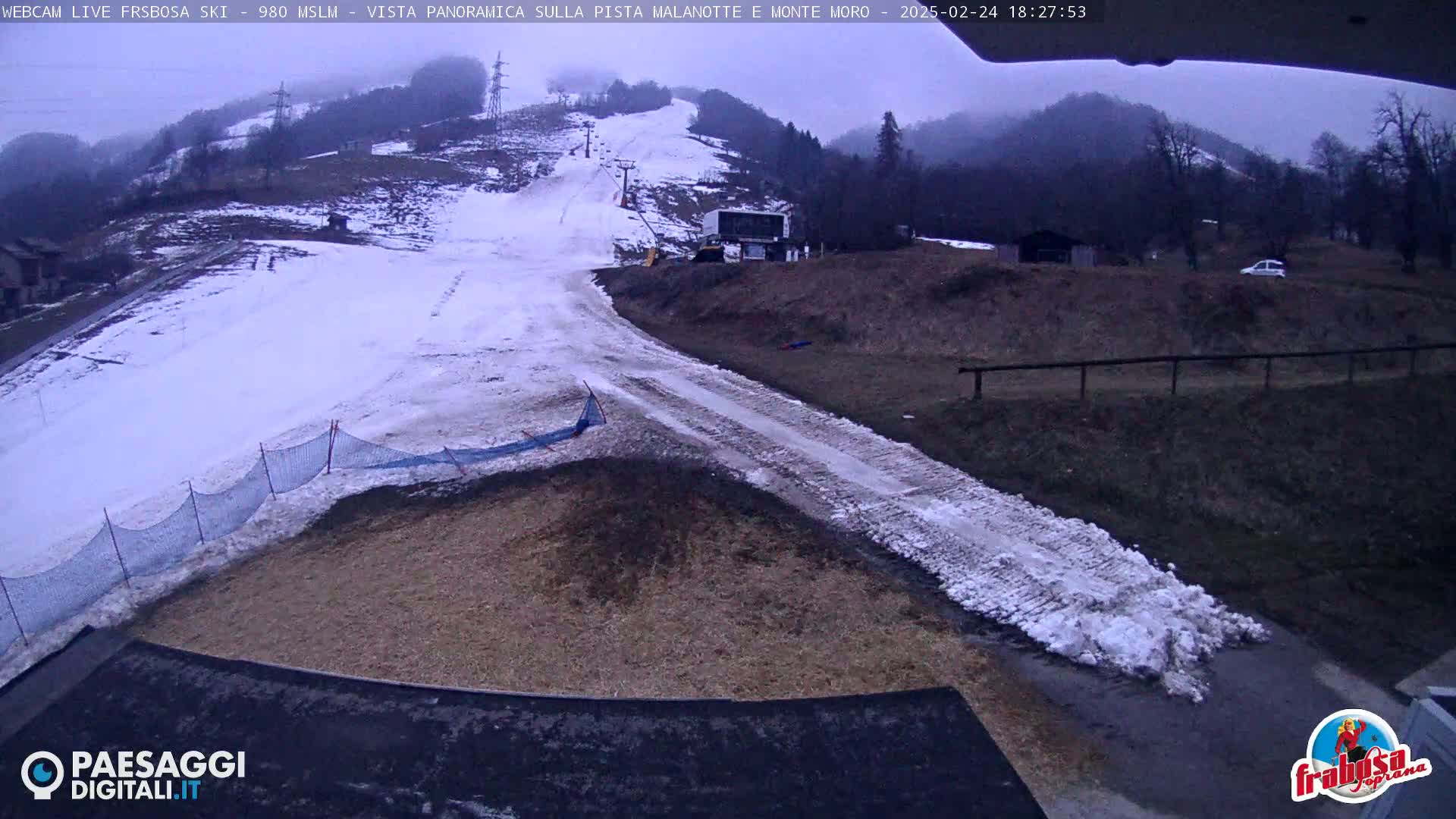 A snow-covered ski slope with a muddy path leading to a small building, viewed under overcast skies.